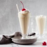 caramelized pineapple milkshake in a glass with whipped cream and cherry and on a stack of plates.