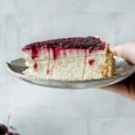 Side view of slice of cherry cheesecake on a plate held in a hand.
