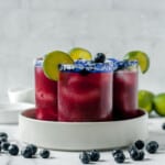 three blueberry margaritas on a tray with blueberries and lime slices.