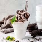 Scoops of mint oreo ice cream in a glass dish with spoon and crumbled oreos on the side.
