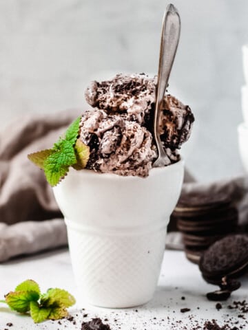Scoops of mint oreo ice cream in a glass dish with spoon and crumbled oreos on the side.