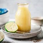 Glass jug of honey lime dressing on a stack of plates with a lime half.